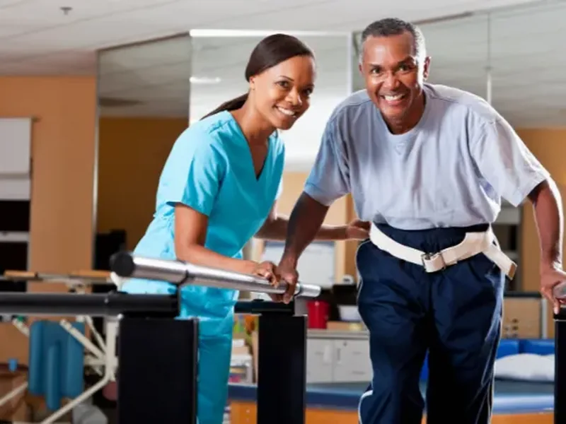 A smiling therapist in light blue scrubs helping a smiling man walk between parallel bars with a gait belt