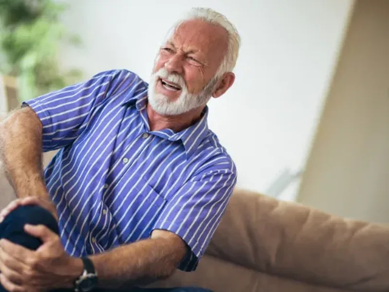 An older man wincing and holding his knee as he sits on a couch