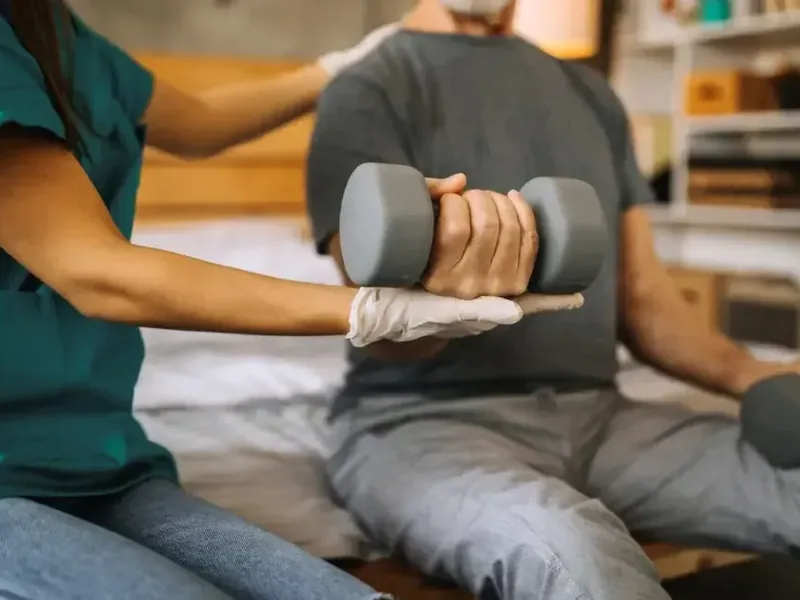 A therapist in green scrubs helping a patient lift a dumbbell during a hand and wrist exercise