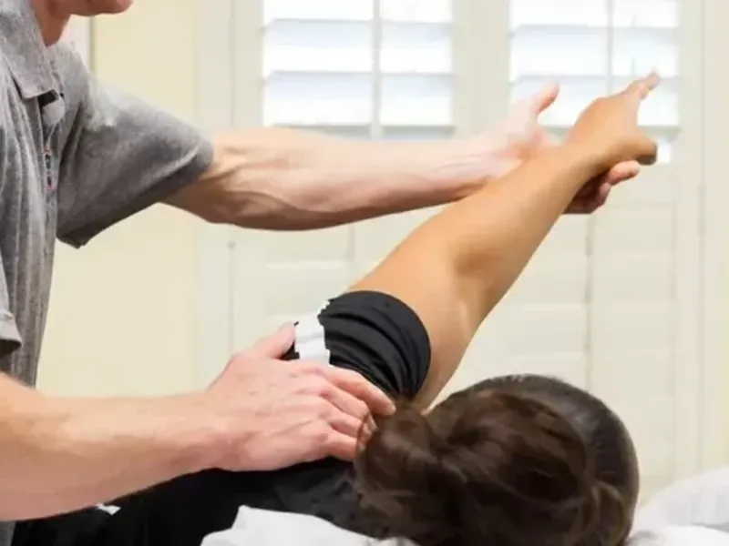 A therapist guiding a woman&rsquo;s raised arm as she lies on a treatment table