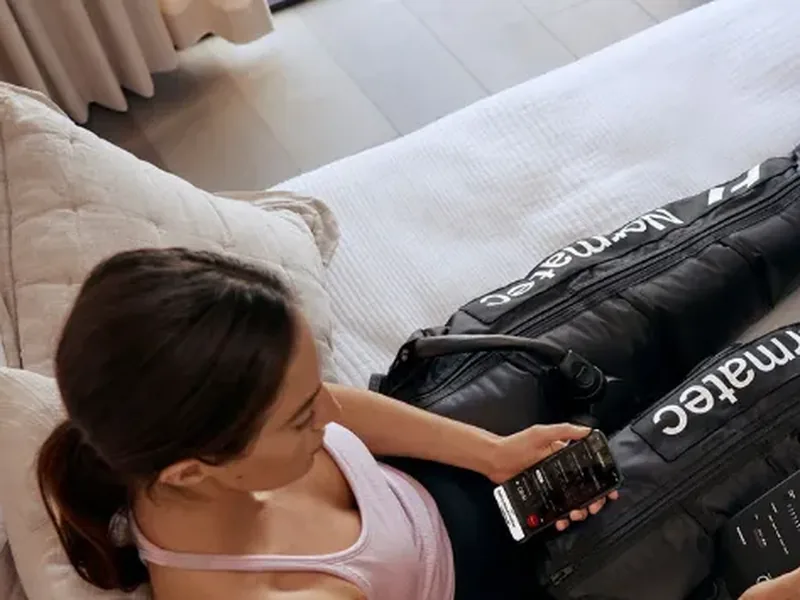 A woman lying back on a bed in Normatec compression boots, looking at her phone