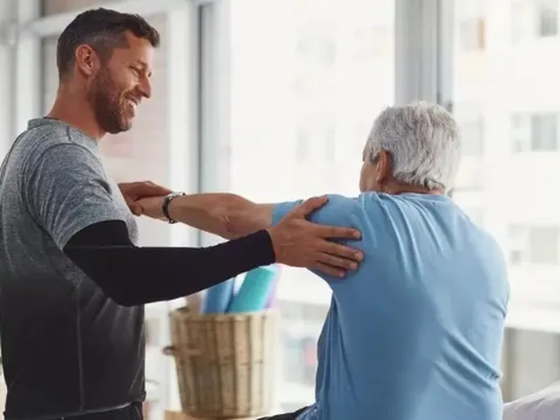 A smiling physical therapist guiding an older man&rsquo;s arm to stretch his shoulder