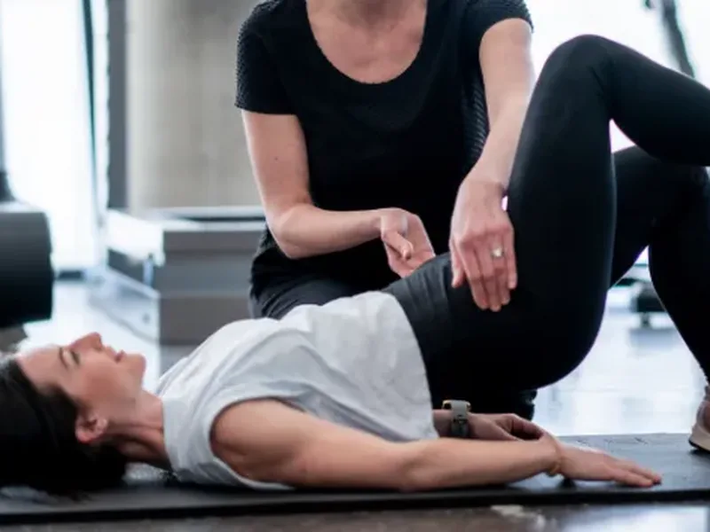 A trainer guiding a woman&rsquo;s bent leg as she lies on a mat in a gym