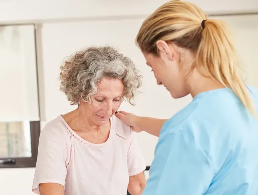 A therapist in light blue scrubs steadying an older woman as she grips a parallel bar during a session